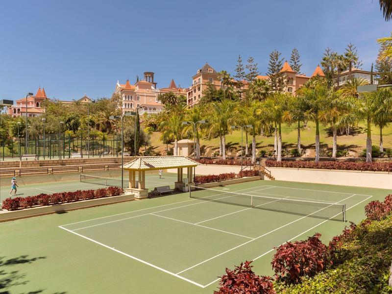 Venkovní tenisový kurt obklopený palmami a tropickou zelení poblíž hotelových budov.