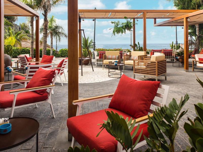 Venkovní posezení s červenými polstrovanými židlemi a stolky obklopené rostlinami a palmami.