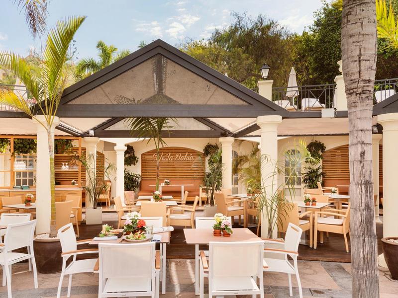 Venkovní jídelna s bílým nábytkem a palmami pod zastřešenou terasou.