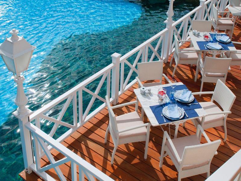 Venkovní jídelní prostor se stoly a židlemi u průzračného modrého bazénu a bílého zábradlí.