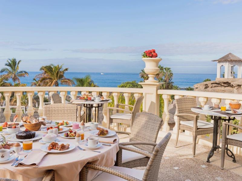 Venkovní terasa s jídelními stoly s výhledem na moře a palmy pod jasnou oblohou.