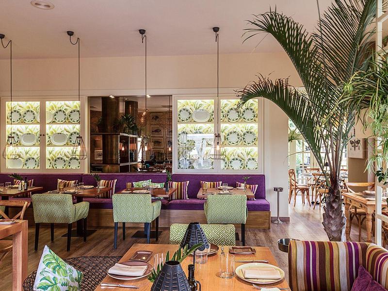 Světlý hotelový jídelní prostor s fialovými pohovkami, zelenými židlemi, dřevěnými stoly a vysokými palmami.