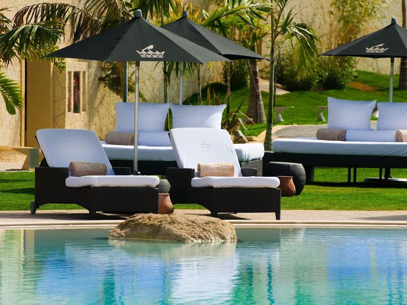 Venkovní lehátka a pohovky s polštáři u čistého bazénu a slunečníky na zahradě.