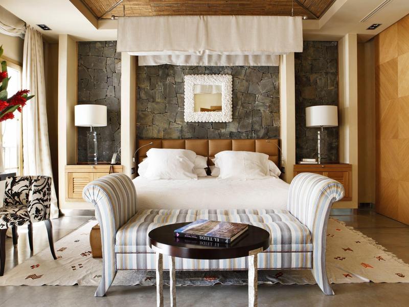 Pohodlný hotelový pokoj s velkou postelí, pruhovanou pohovkou, lampami a květinovým křeslem.