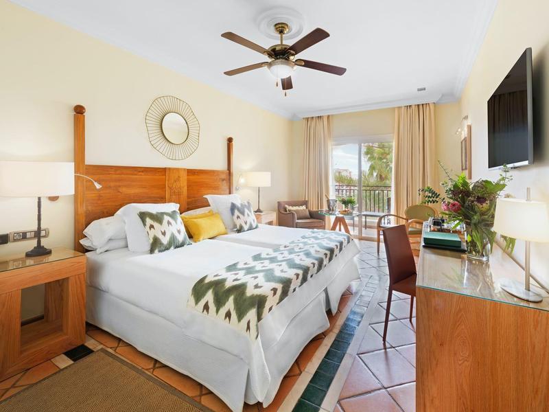 Světý hotelový pokoj s manželskou postelí, dřevěným nábytkem, stropním ventilátorem a výhledem na balkon.