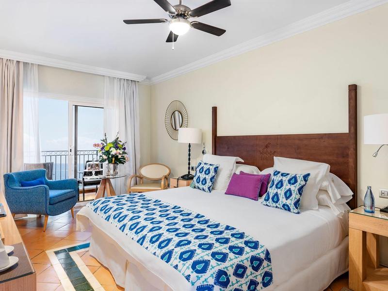 Světý hotelový pokoj s manželskou postelí, modrým křeslem a balkonem s výhledem na oceán.