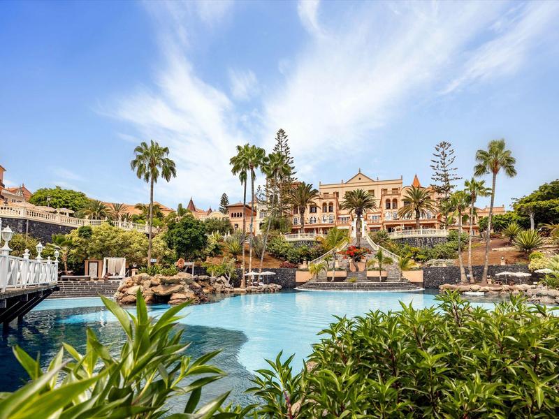 Luxusní hotel s tropickými palmami a velkým bazénem pod modrou oblohou.