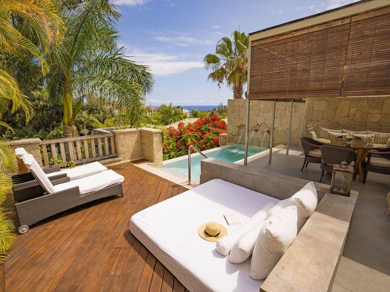 Venkovní terasa s lehátky, jídelním stolem, malým bazénem a tropickými rostlinami pod modrou oblohou.