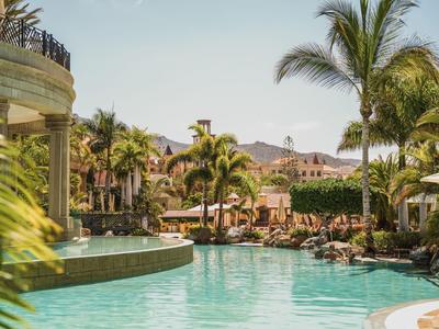 Grande piscine d'hôtel avec palmiers et transats sous un ciel ensoleillé et montagnes en arrière-plan.