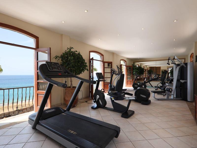 Helles Fitnessstudio mit Laufband, Fahrradergometer, Blick aufs Meer und großen Fenstern.