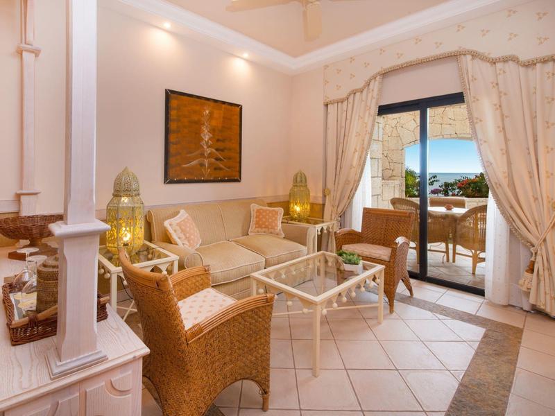 Helles Wohnzimmer mit Korbmöbeln, Glas-Couchtisch und Blick auf Terrasse und Meer durch große Fenster.