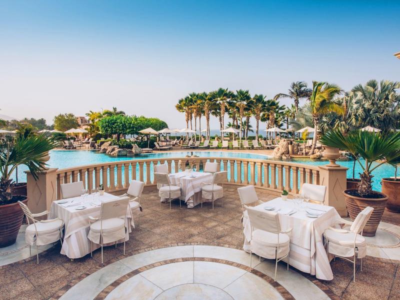 Terrasse mit weißen Tischen, Stühlen, Palmen und Blick auf Pool und Meer bei klarem Himmel.