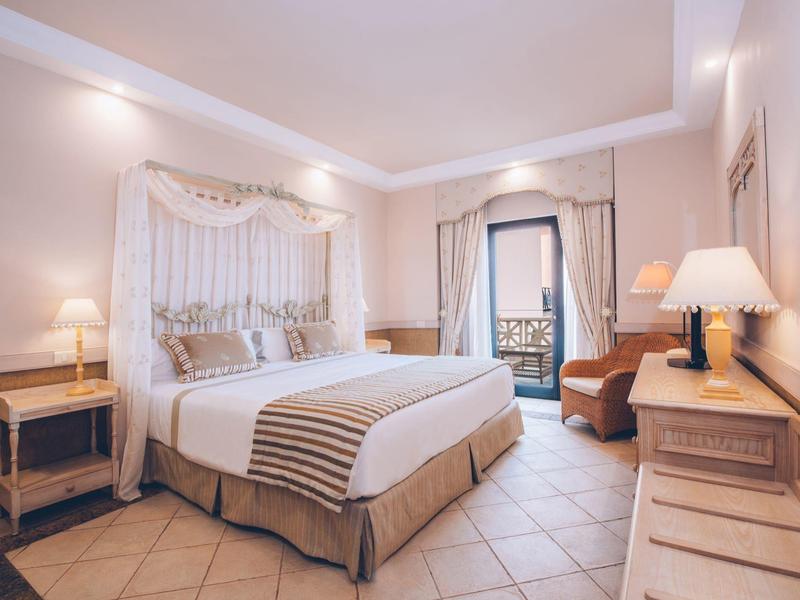 Helles Schlafzimmer mit großem Bett, beiger Bettwäsche, Sesseln, Nachttischen und Balkon mit Stuhl.