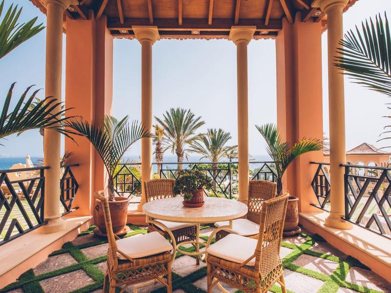 Terrassenbereich mit runder Tischgruppe aus Holz, Pflanzen und orangefarbenen Säulen, Palmenblick.