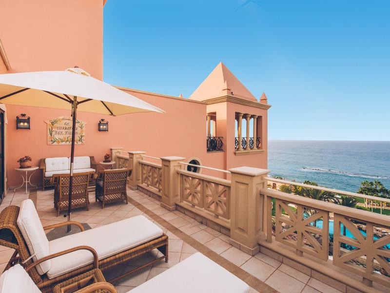 Balkon mit Stühlen, Tisch, Sonnenschirm und Meerblick unter klarem blauem Himmel.