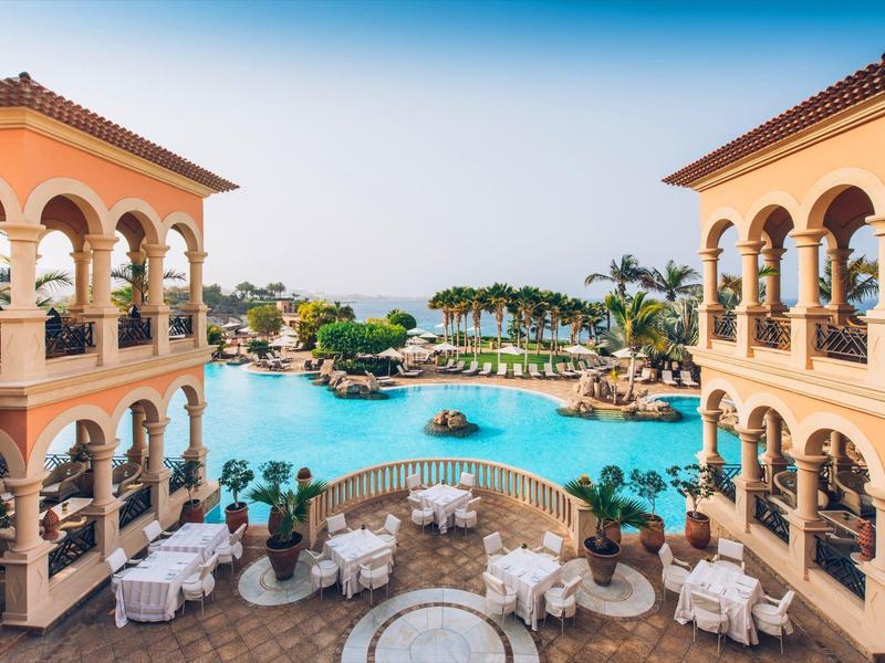 Großer Pool mit Palmen, umgeben von mediterranen Gebäuden und Sitzbereichen im Freien.