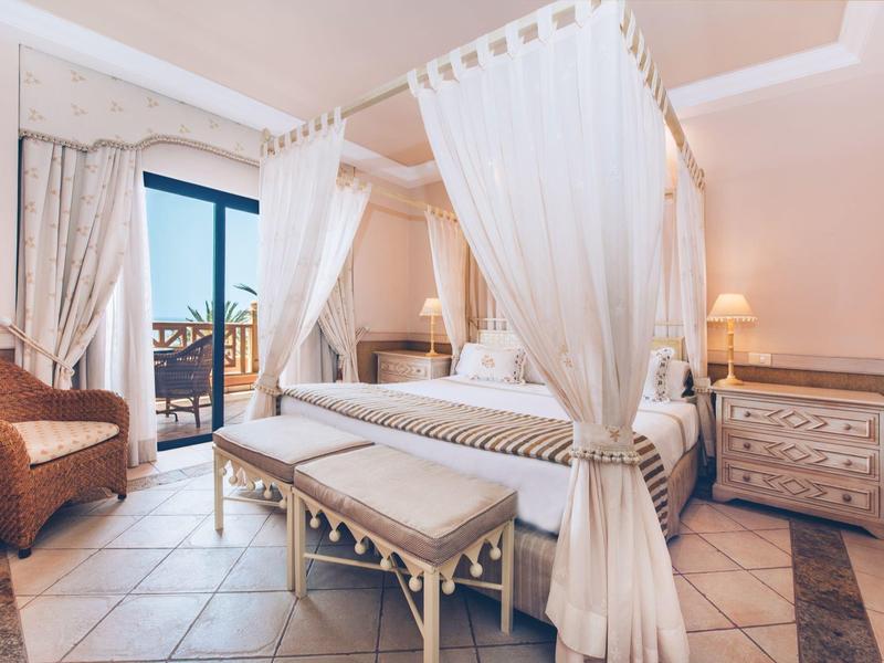 Gemütliches Schlafzimmer mit Himmelbett, Sitzbank, Rattanstuhl und Balkon mit Blick nach draußen.