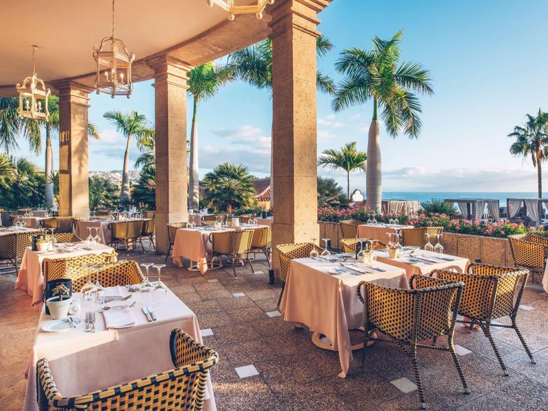Offenes Restaurant mit gedeckten Tischen, Säulen und Palmen mit Blick auf das Meer im Hintergrund.