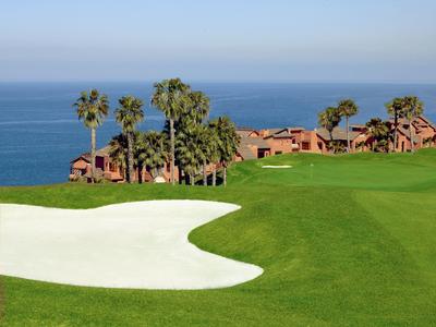 Golfplatz mit grünem Rasen, Sandbunker, Palmen und Häusern am Meer im Hintergrund.