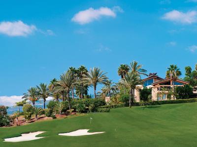 Grünes Golfplatzfeld mit Sandfallen, Palmen und einem Haus im Hintergrund unter blauem Himmel.
