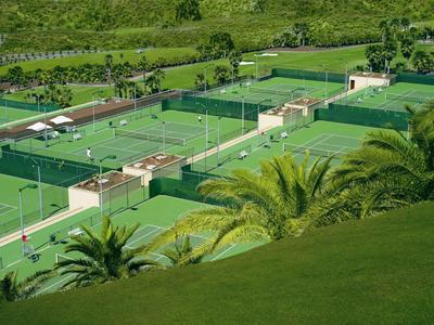 Mehrere grüne Tennisplätze mit Netzen und Umzäunungen, umgeben von Palmen und Hügeln.