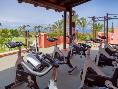 Moderner Fitnessbereich mit mehreren Ergometer-Fahrrädern unter überdachtem Bereich, Palmen im Hintergrund.