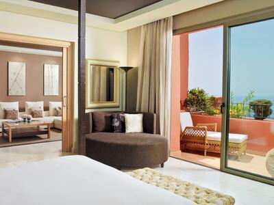 Helles Schlafzimmer mit großem Fenster, gemütlicher Sitzecke und Blick auf Terrasse mit Meer.