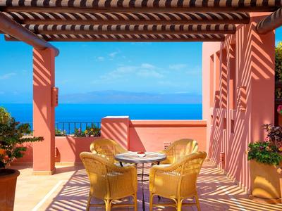 Terrasse mit gelben Sesseln, Tisch, pinken Wänden und blauem Meer im Hintergrund bei Sonnenschein.