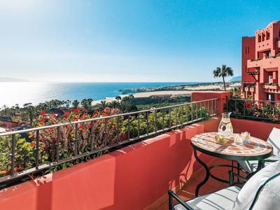 Terrassenbereich mit Glas-Tisch, Stühlen und Blick auf Meer, Palmen und Gebäude in rötlicher Farbe.
