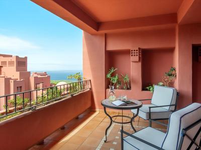 Balkon mit Stühlen und Tisch, Blick auf Meer und rosa Gebäude bei blauem Himmel.