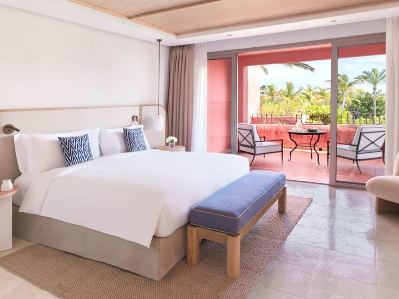 Lichte hotelkamer met groot bed, bankje, fauteuil en toegang tot terras met uitzicht op de tuin.