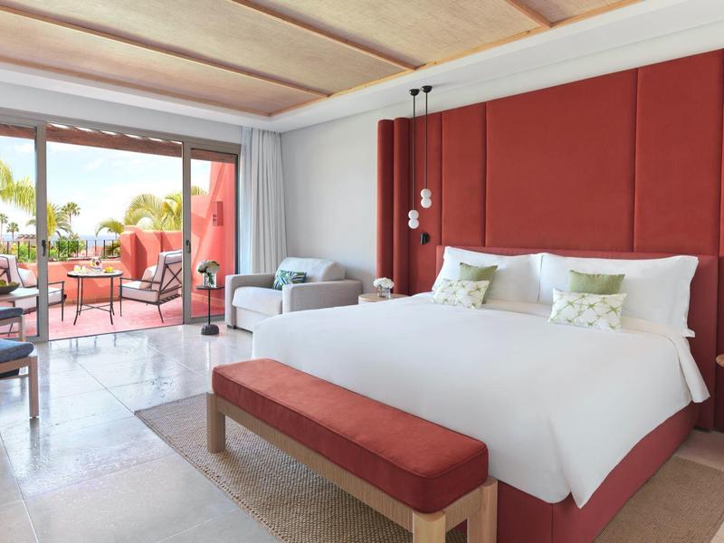 Moderne hotelkamer met groot bed, rode accenten en terras met uitzicht op zee.