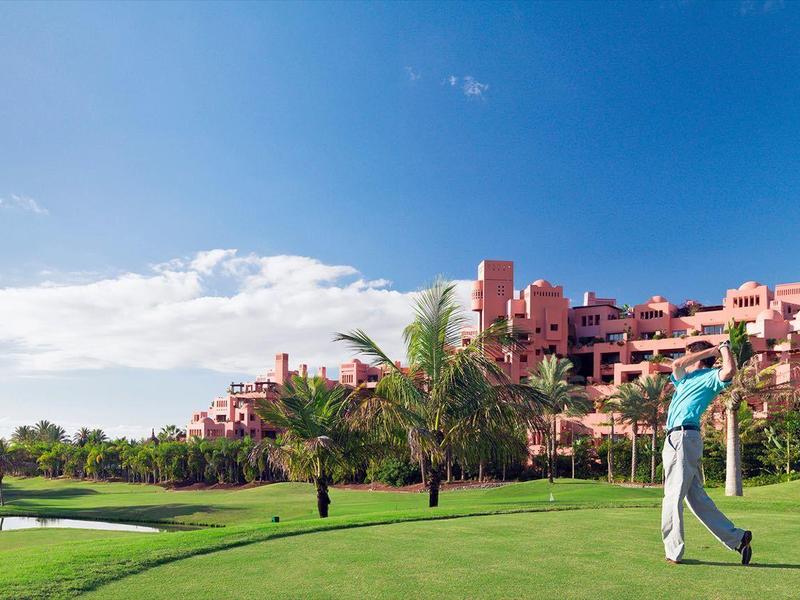 Golfer staat op goed onderhouden gras voor hotel met palmbomen en blauwe lucht.