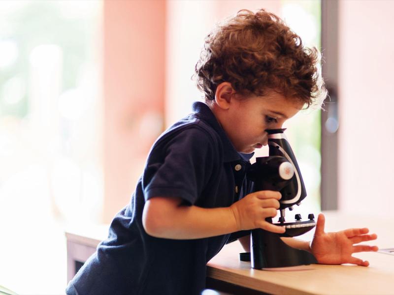 Klein kind kijkt nieuwsgierig door een microscoop op een tafel in een lichte kamer.