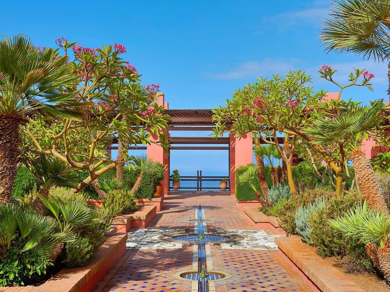 Een betegeld pad leidt door tropische planten naar een poort met uitzicht op de oceaan.