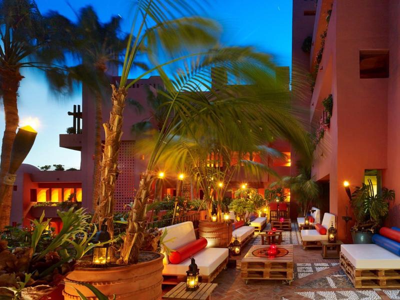 Gezellige buitenlounge met banken, kussens, palmbomen en warme verlichting bij schemering.