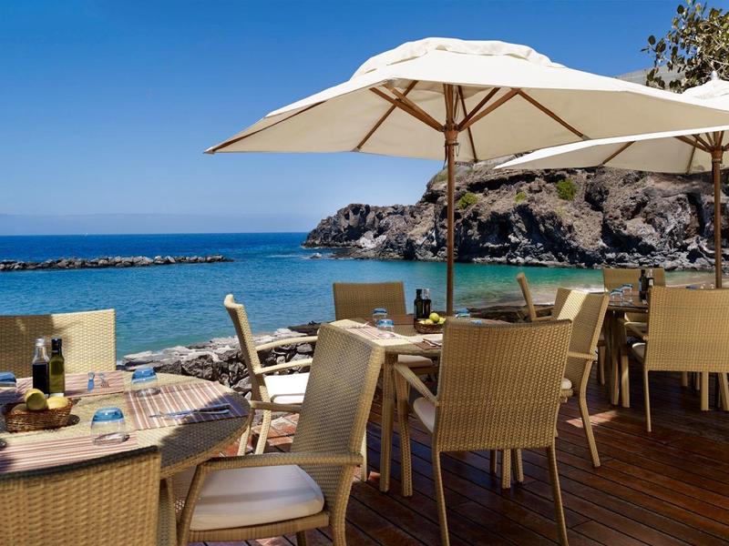 Strandrestaurant met tafels, stoelen en grote parasols met uitzicht op zee en rotskust.