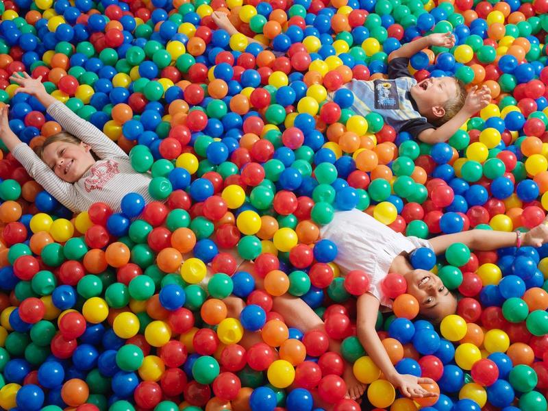 Drie kinderen ontspannen en spelen in een kleurrijk ballenbad met rode, blauwe, gele en groene ballen.