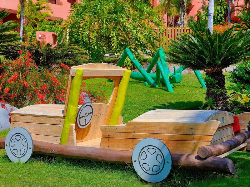 Een man ligt op het gras naast een houten autostoel in een tropische tuin.