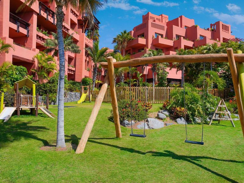Speeltuin met schommels en grasveld voor een meerlaags hotelgebouw op een zonnige dag.