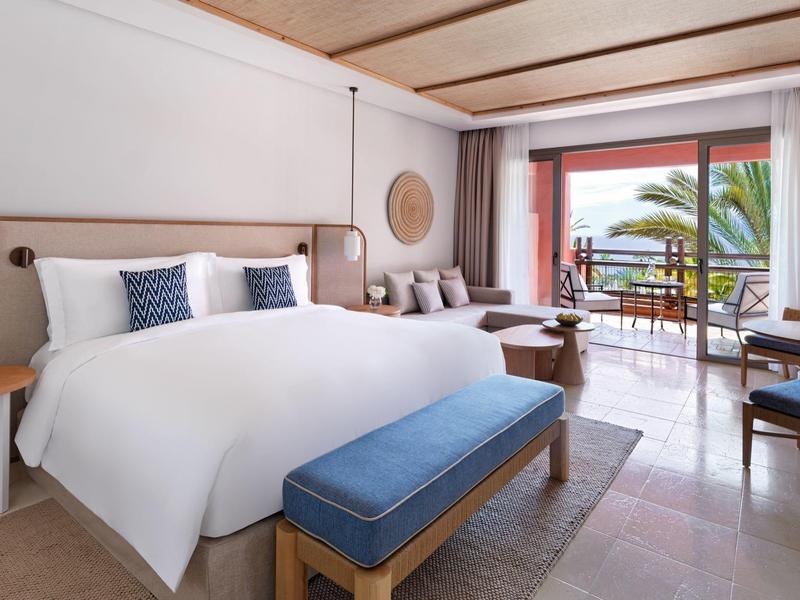 Modern hotelkamer met groot bed, blauwe accenten en balkon met uitzicht op zee.