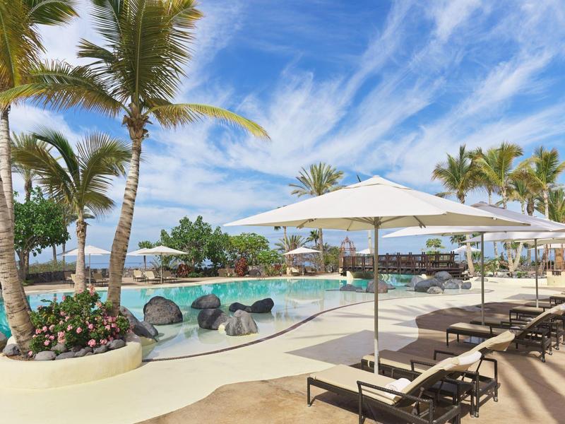 Moderne zwembadruimte met ligstoelen onder parasols en palmbomen onder een blauwe lucht.