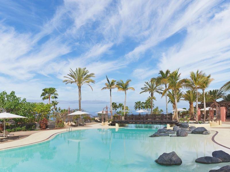 Een rustig hotelzwembad met palmbomen, parasols en een blauwe lucht.