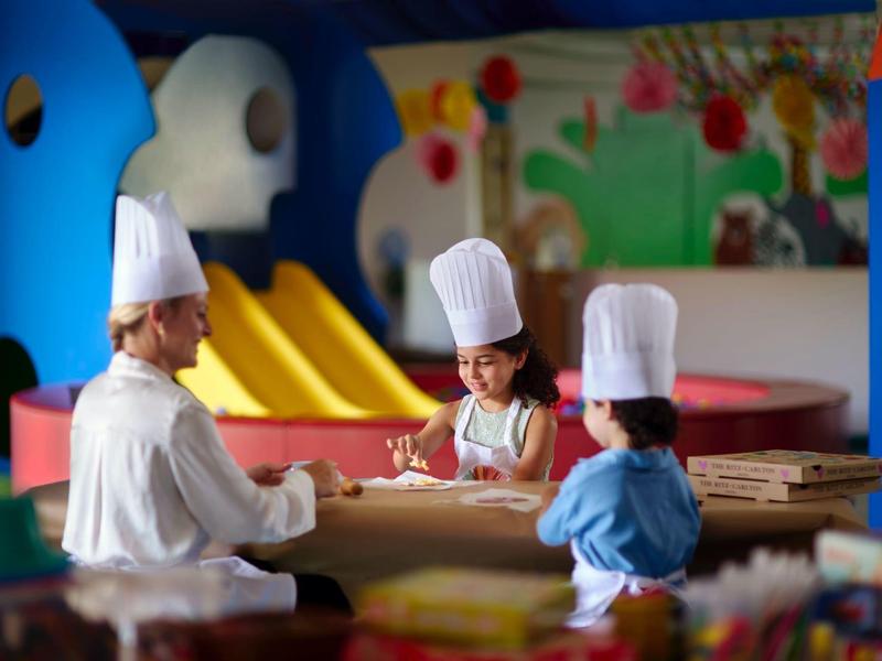 Een kok leert twee kinderen met kokshelmen in een kleurrijke speelruimte.