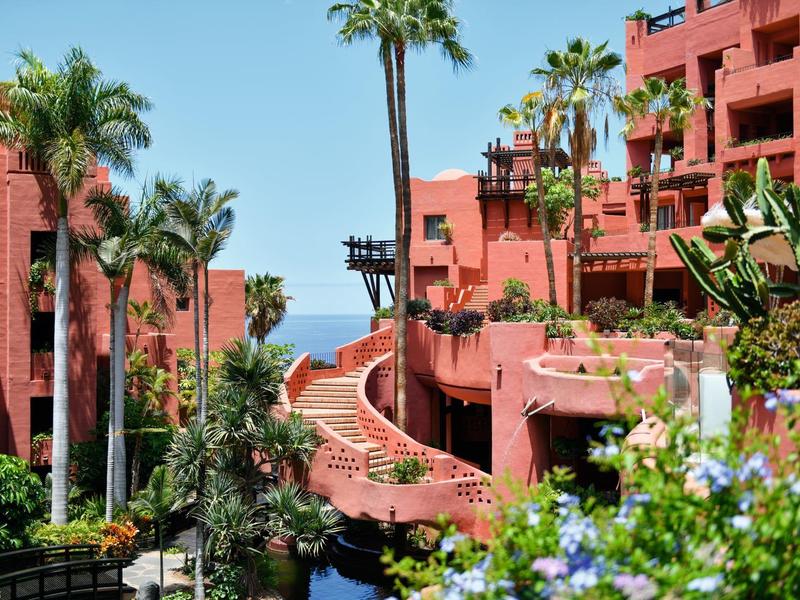 Hotelcomplex met rode gebouwen, palmbomen en een pad dat naar zee leidt.