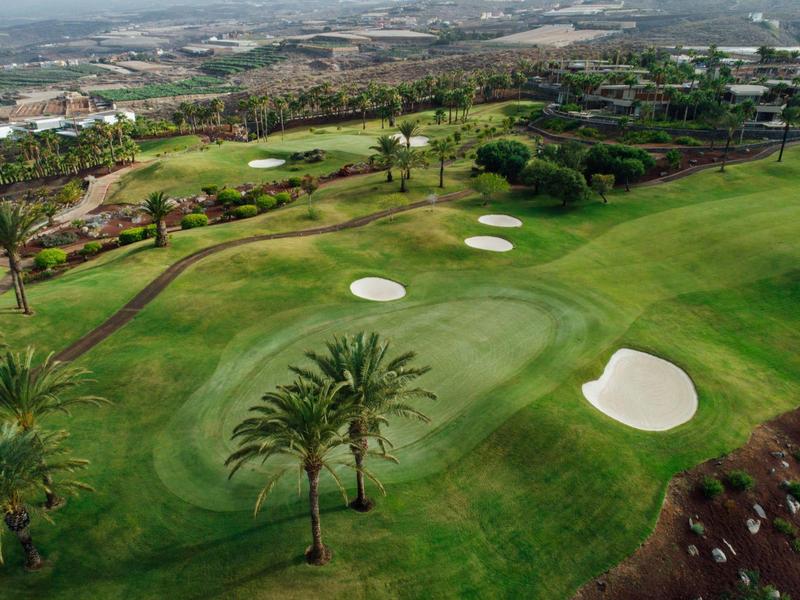 Parcours de golf vert avec bunkers de sable et palmiers devant une vue urbaine lointaine.