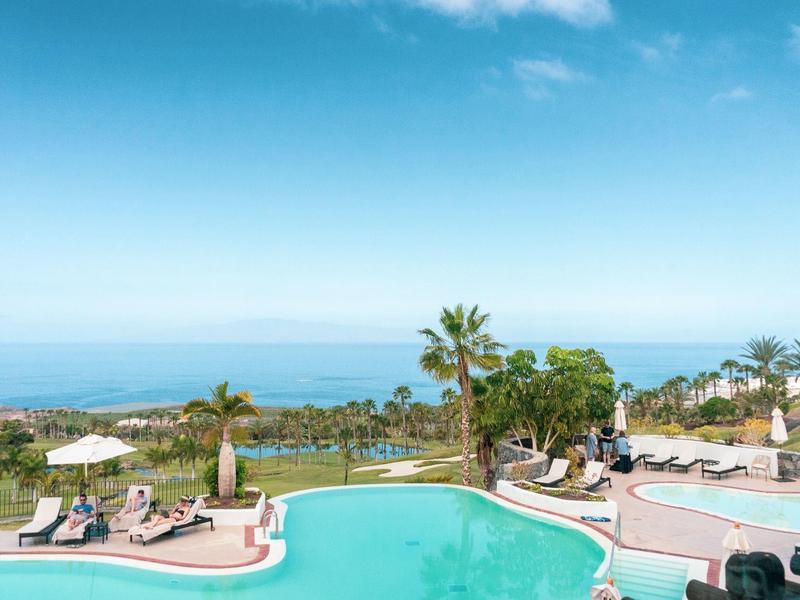 Vue d'une piscine avec des palmiers et la mer en arrière-plan sous un ciel bleu.