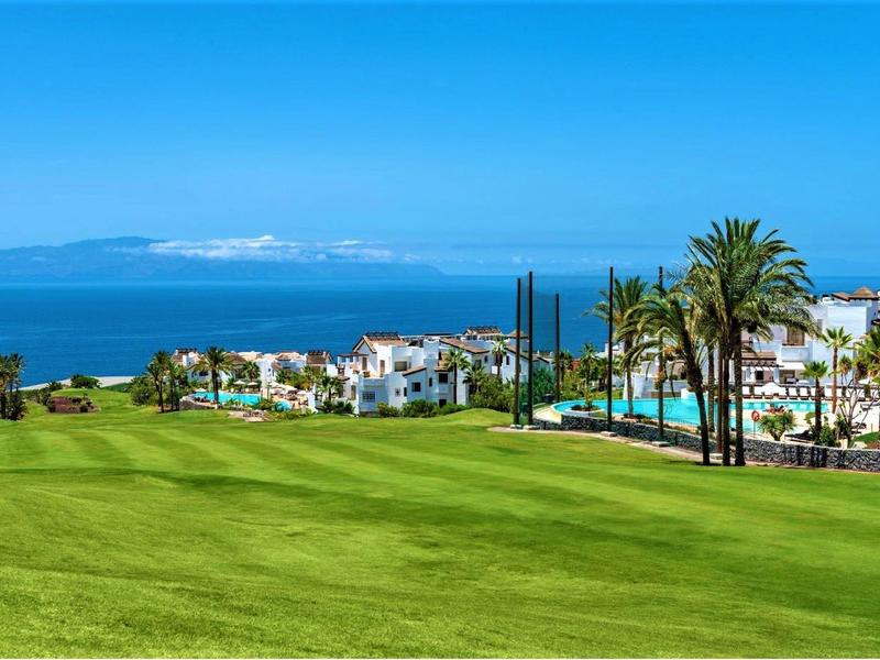 Pelouse verte avec bâtiments blancs, palmiers et mer en arrière-plan sous un ciel clair.
