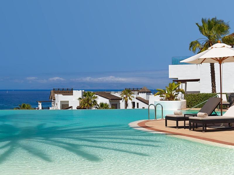 Piscine à débordement avec chaises longues et parasol, vue sur la mer et les hôtels.