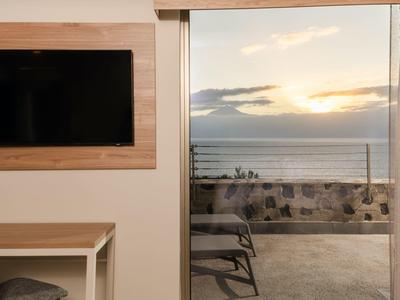 View of a balcony with chair and sea view at sunset, TV and desk inside the room.